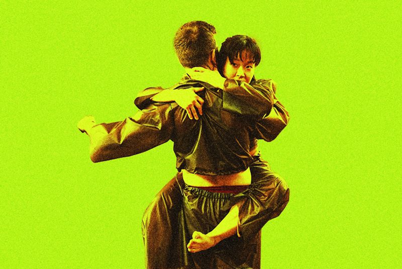 A dancer is shown from the back, wearing a dark, shiny tracksuit, while being embraced by a second dancer whose face is visible over their shoulder. The image is set against a vibrant, solid lime-green background with a slightly grainy, high-contrast texture.