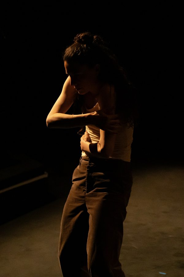 A woman with long hair, wearing a white tank top and dark trousers, is illuminated by low light as she wraps her arms around herself and curves her back.