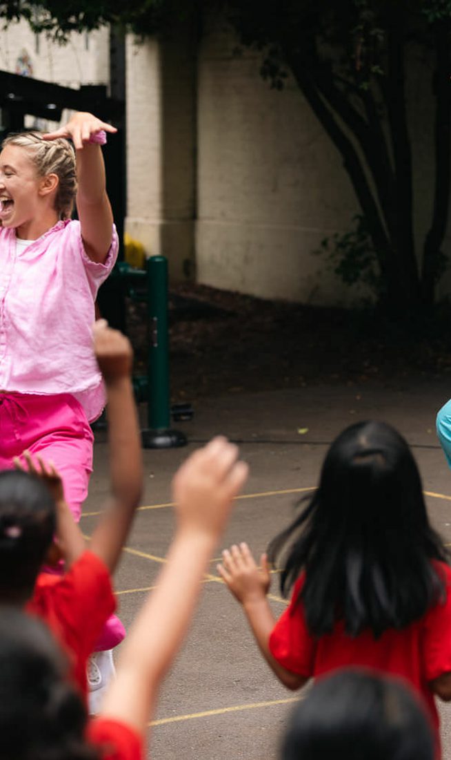 Two dancers - one in pink and one in blue - dance energetically in a school playground whilst children wave their arms and jump in front of them