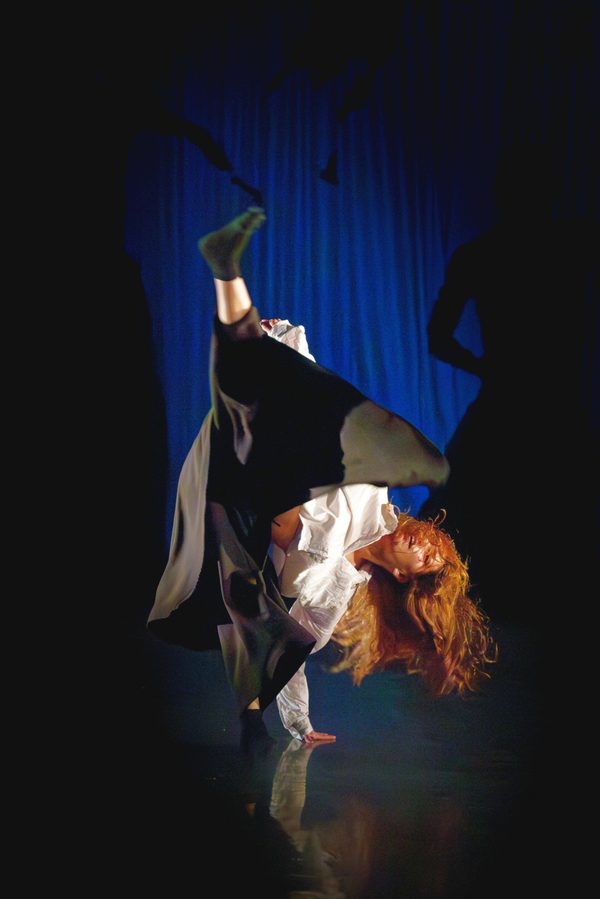 A dancer with long, flowing hair performs a dynamic floor move onstage, balancing on one hand with one leg extended upward. They wear a white top and dark, loose trousers, illuminated against a deep blue backdrop with shadowy figures in the background.