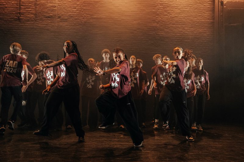 A group of children in matching burgundy T-shirts with white logos, black trousers, and sneakers dancing on stage.